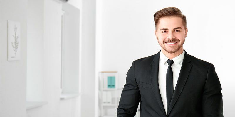 Hombre joven con barba de traje negro, camisa blanca y corbata sonriendo