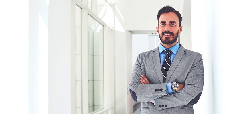 Hombre de barba con traje gris, camisa azul y corbata,  sonriendo con los brazos cruzados