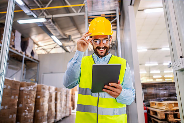 ingeniero de mediana edad de barba y anteojos, con casco y chaleco de seguridad amarillo sonriendo mientras revisa los datos de su tablet