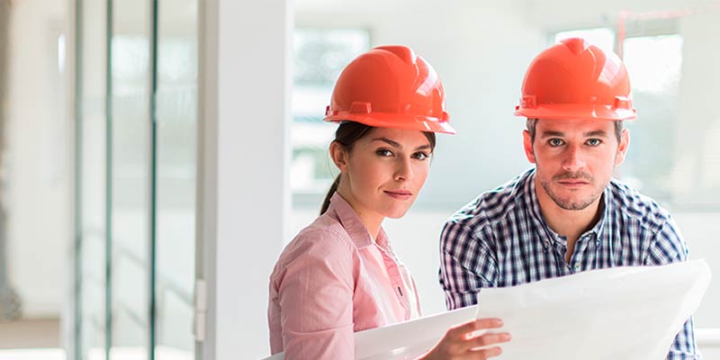 Una pareja de  ingenieros jovenes con casco rojo revisando un plano de una edificación