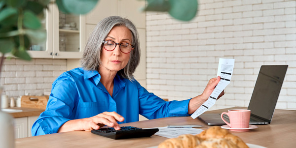 mujer de la tercera edad con factura en mano,  realizando cuentas en una calculadora frente a su laptop