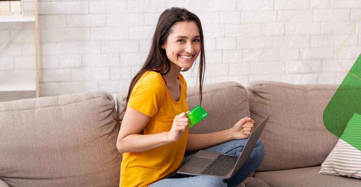 Mujer feliz sentada en el sofá con una laptop y una tarjeta de crédito de Banco Popular en la mano