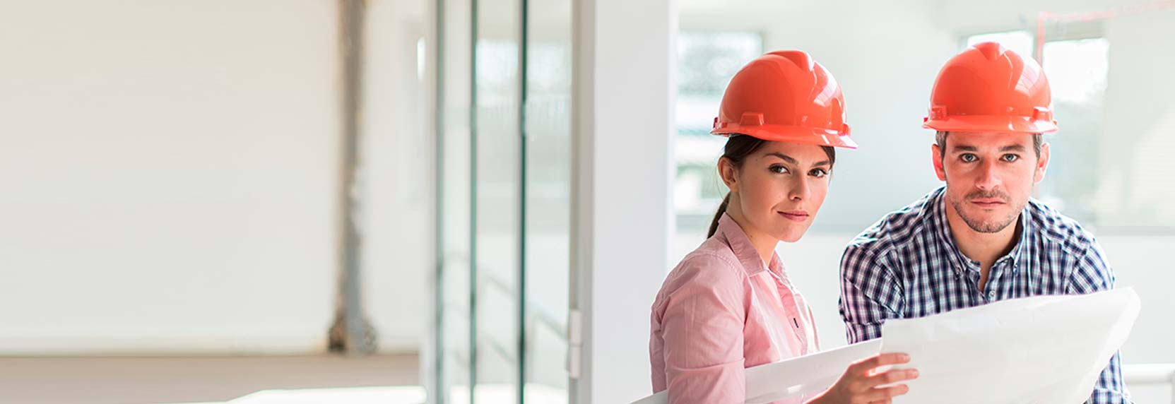 Una pareja de  ingenieros jovenes con casco rojo revisando un plano de una edificación